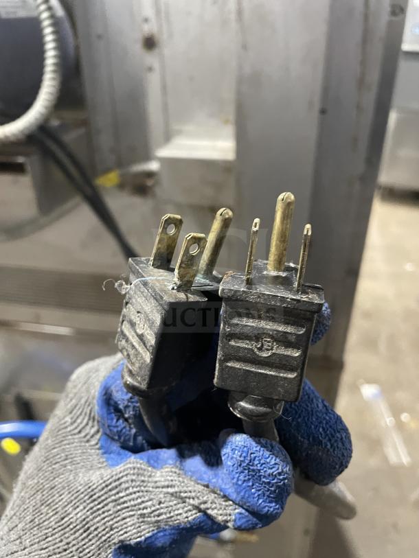 Two electrical plugs held by a gloved hand, in front of a metallic background.