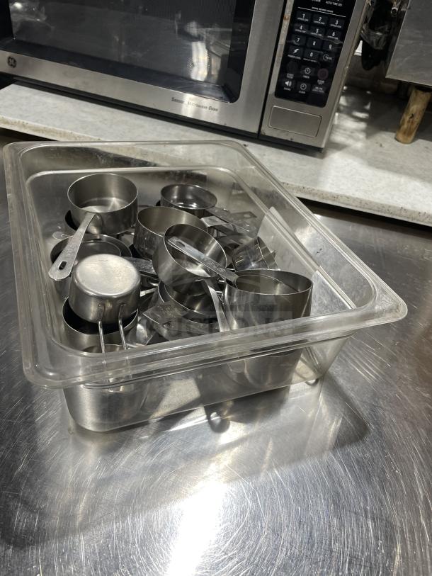 Commercial stainless steel measuring cups in a transparent container on a metal surface. Good condition.
