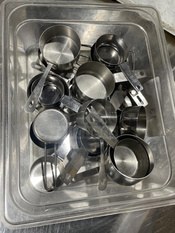 Stainless steel measuring cups in a plastic container, various sizes, marked with measurements, in good condition.