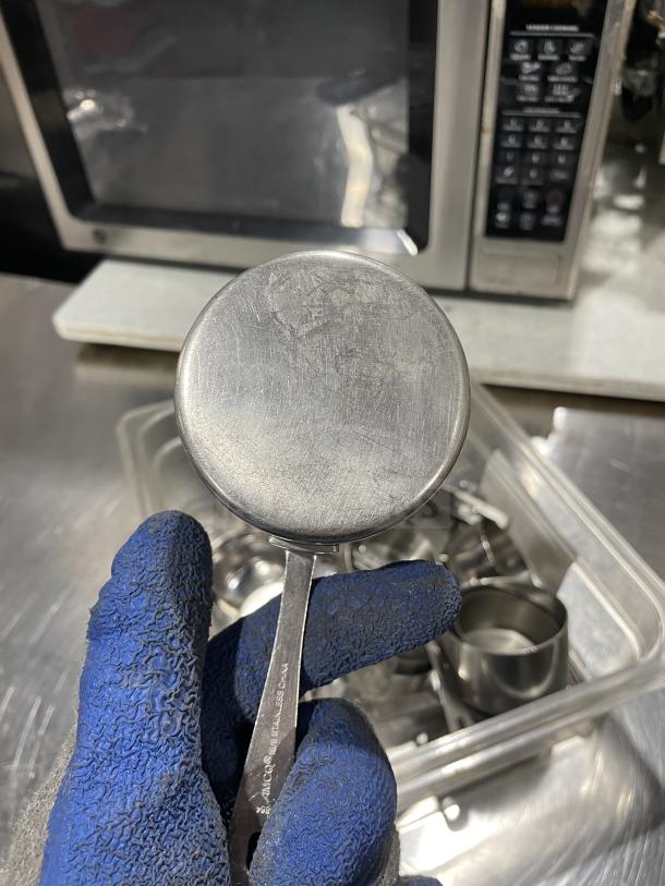Commercial stainless steel measuring cups, lightly used, held in gloved hand. Engraved markings visible on handle.