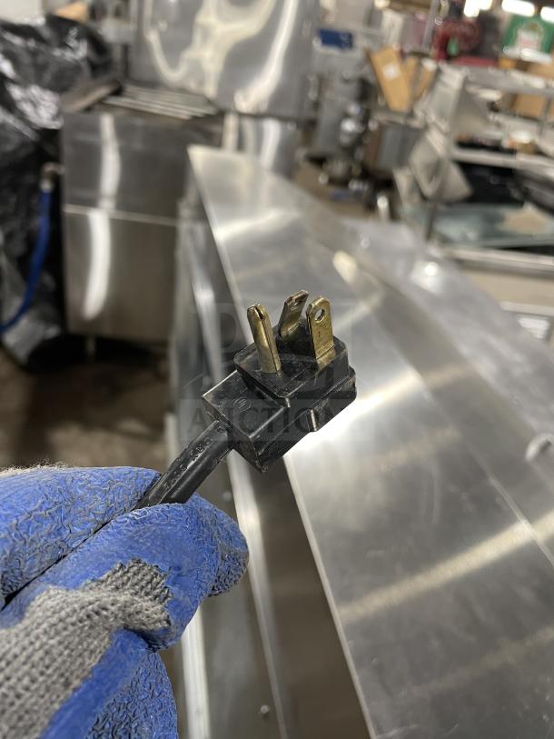 Gloved hand holding a three-prong electrical plug near a stainless steel commercial prep table.