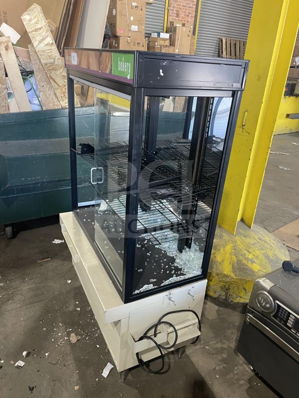Commercial bakery display case with glass panels, metal frame, and shelves; visible damage includes scattered broken glass.