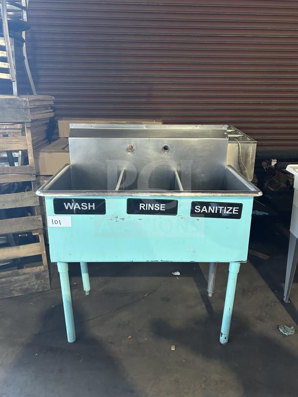 Commercial teal 3-compartment stainless steel bar sink with labels "Wash," "Rinse," "Sanitize." On sturdy legs.