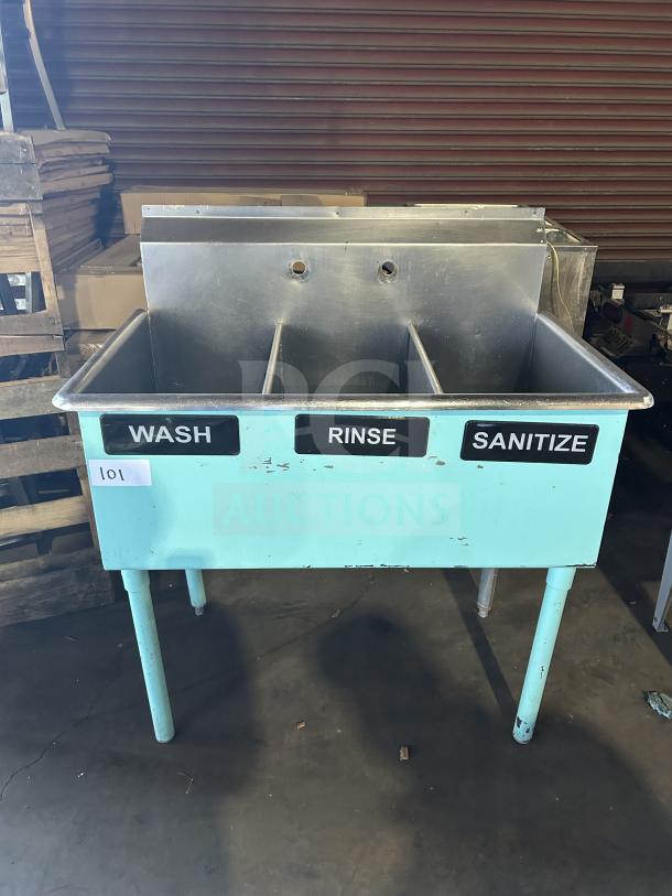 Commercial stainless steel teal 3-compartment bar sink with "Wash," "Rinse," "Sanitize" labels. On legs. Good condition.