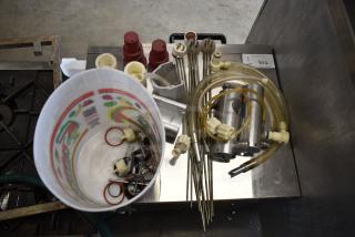 92 various ice cream machine parts, including handles, hoses, and metal cylinders, in a mixed condition on a metallic surface.