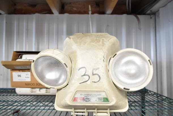 Emergency light with dual lamps, beige body, marked "35," on a metal shelf. Additional boxed items around it.