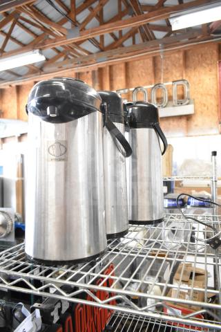 Three stainless steel air pots with black lids and handles, labeled "Service Ideas," on a metal shelf. Good condition.
