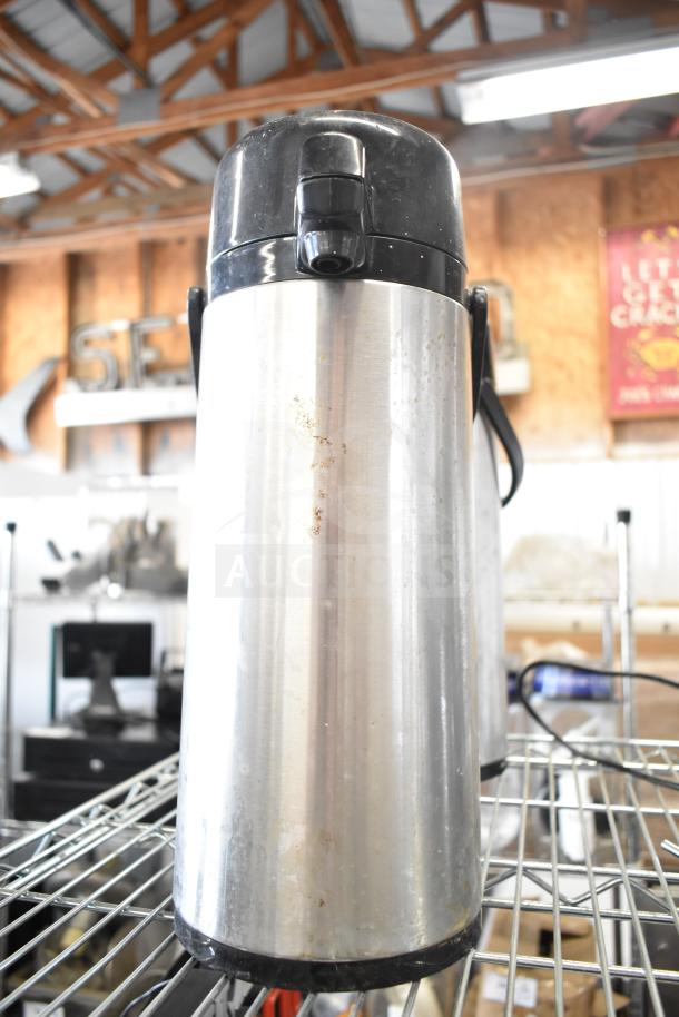 Three stainless steel air pots with black lids on a metal shelf, showing signs of wear. Ideal for hot beverage service.