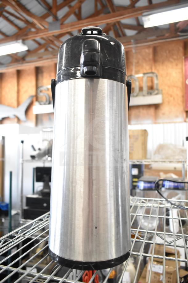 Stainless steel air pot with black lid and handle, in good condition, on metal shelf in a warehouse setting.