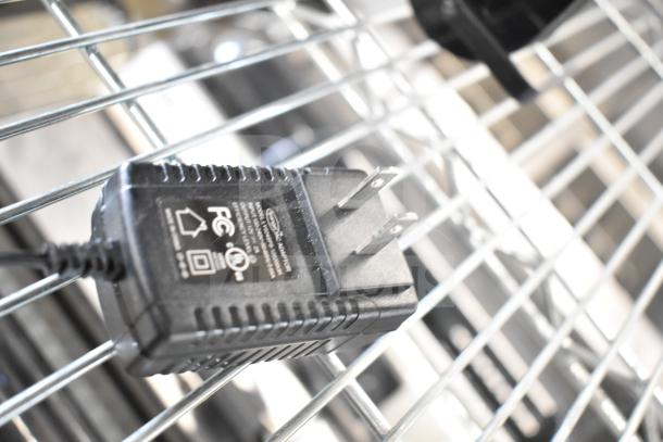 Power adapter with visible voltage specifications and FCC certification, resting on a metal rack.