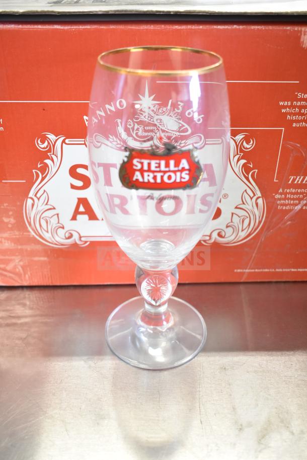 Stella Artois beer glass with gold rim, branded logo, clear stem, and decorative base, in front of branded box.