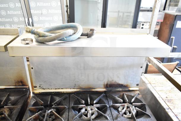 Commercial gas-powered 6 burner range with oven, over shelf, and back splash. Features visible wear. Ideal for heavy-duty use.