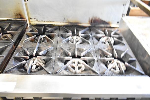 Commercial gas-powered 6-burner range with oven, over shelf, and backsplash. Visible wear, suitable for professional kitchens.