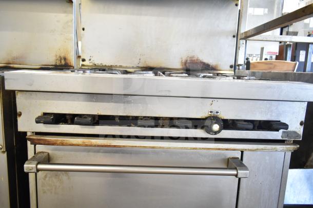 Commercial gas-powered 6-burner range with oven, over shelf, and backsplash. Visible wear, stainless steel build.