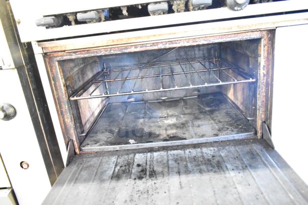 Commercial gas-powered 6-burner range with oven, over shelf, and backsplash, showing signs of use. Oven door open, interior visible.