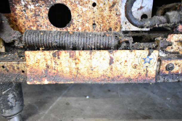 Rusted component of a commercial 6-burner gas range with visible wear and tarnish. Includes spring coil and faded label.