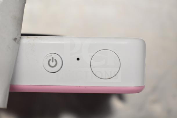 Close-up of a white camera with a pink trim, showing power button. Condition appears used, with slight dirt visible.