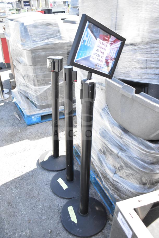 Three black stanchions with retractable belts, used condition, displayed outdoors. Labels on bases indicate auction details.