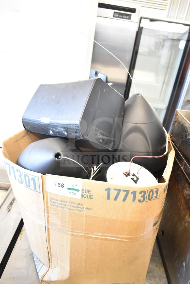 Eight speakers including one QSC AD-S6T and seven Crestron SAROS PD6T-B-T-EACH in a cardboard box, showing moderate wear.