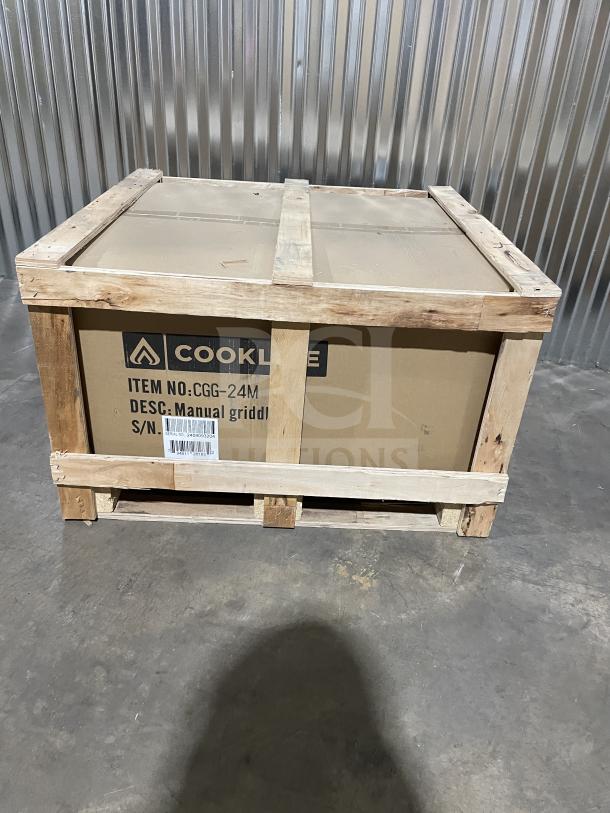 Brand new Cookline CGG-24M gas griddle in crate, features manual controls, stainless steel, back and side splash, on legs.