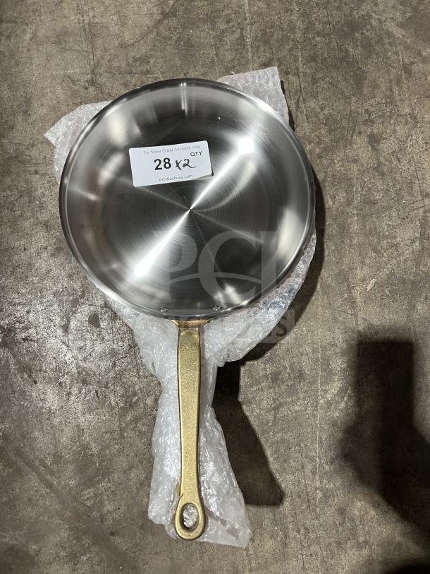Brand new commercial stainless steel frying pan with brass handle, shown with label "28 x2," on bubble wrap.