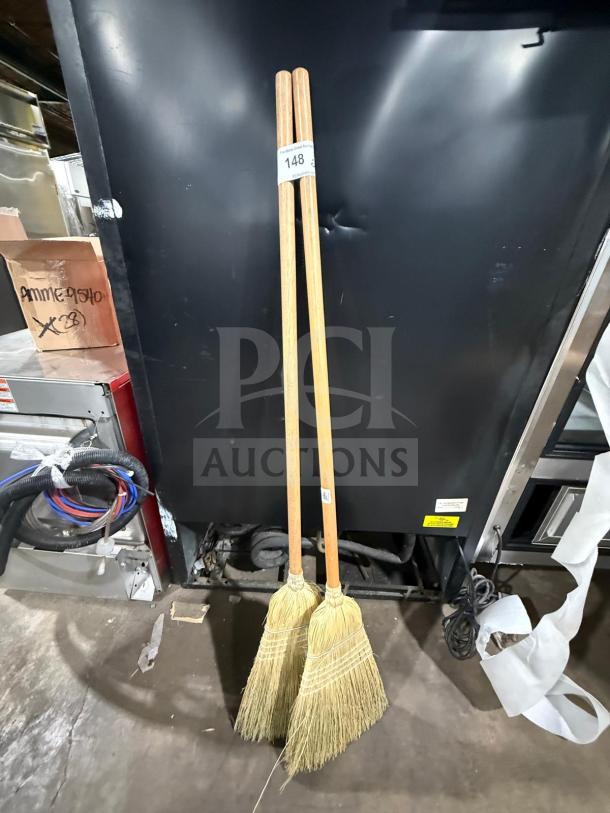 Two new wooden corn brooms with natural bristles, standing upright. Auction tag marked "148".