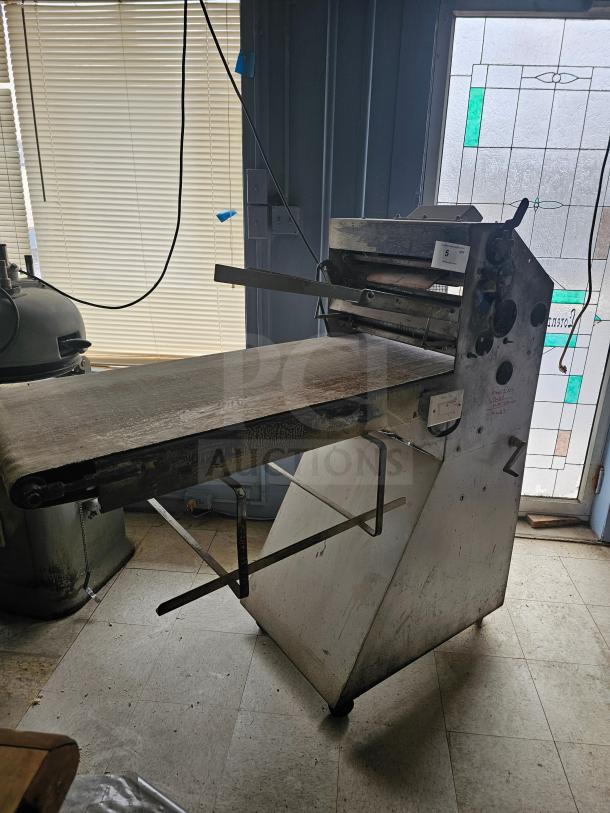 Commercial dough sheeter with metal frame and conveyor, showing some signs of wear. Equipped with control knobs and side levers.