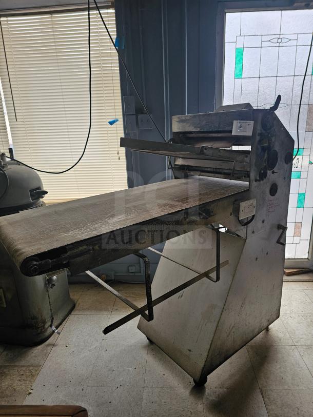 Commercial dough sheeter with metal frame and conveyor belt, showing signs of wear. Suitable for bakery operations.