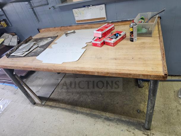 Wood top work table with sturdy metal legs. Appears in used condition with some surface wear. Items on top include boxes, papers, and spray can. Measures approximately 6 feet long. Ideal for workshop or garage use.