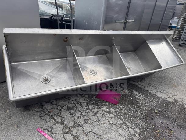 Stainless steel 3 bay sink, industrial-grade, shows signs of use. Each compartment includes a drain. Markings indicate wash, rinse.