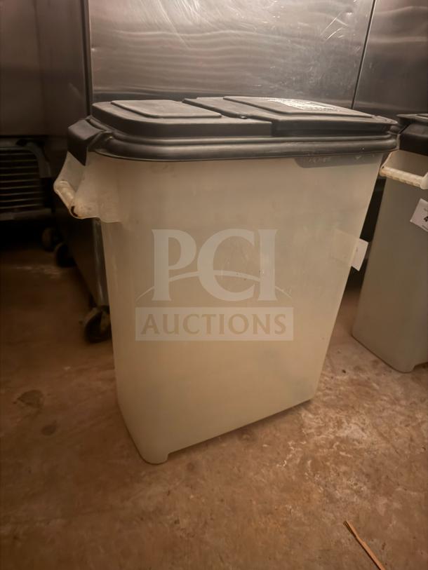 Large translucent storage food container with black lid, industrial setting. Sturdy plastic, moderate wear. Ideal for bulk storage.