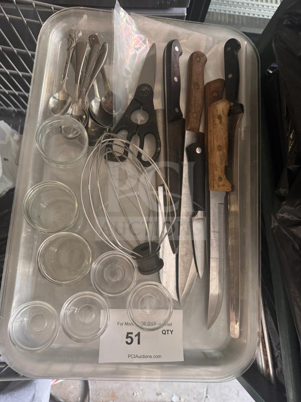 Assorted kitchenware including knives, spoons, glass ramekins, whisk, and shears in used condition in a plastic tray.