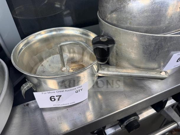 Vintage aluminum food mill with crank handle, perforated bottom, and side hook for stabilization. Auction label with lot number 67 visible.