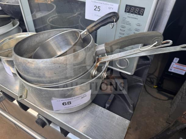 Stacked aluminum saucepans with black handles, showing wear. Visible auction tags indicate items 25 and 68.