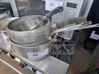 Stacked aluminum saucepans with black handles, showing wear. Visible auction tags indicate items 25 and 68.