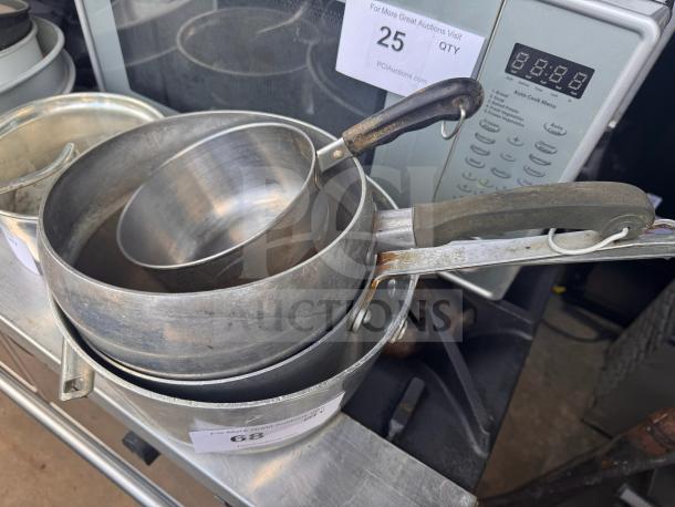 Stacked saucepans with metal handles, showing signs of wear; lot tag "68" attached, sitting on a metal surface.