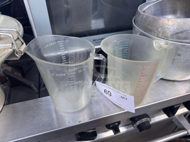 Lot of 2 clear plastic measuring cups with red and white measurement markings, in used condition, displayed on a metal surface.