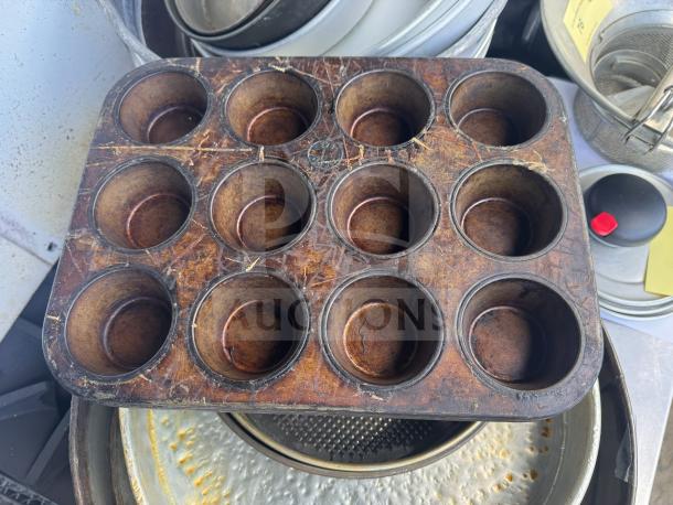Old aluminum muffin pan with 12 cups, showing signs of wear and rust. Distressed surface, likely vintage.