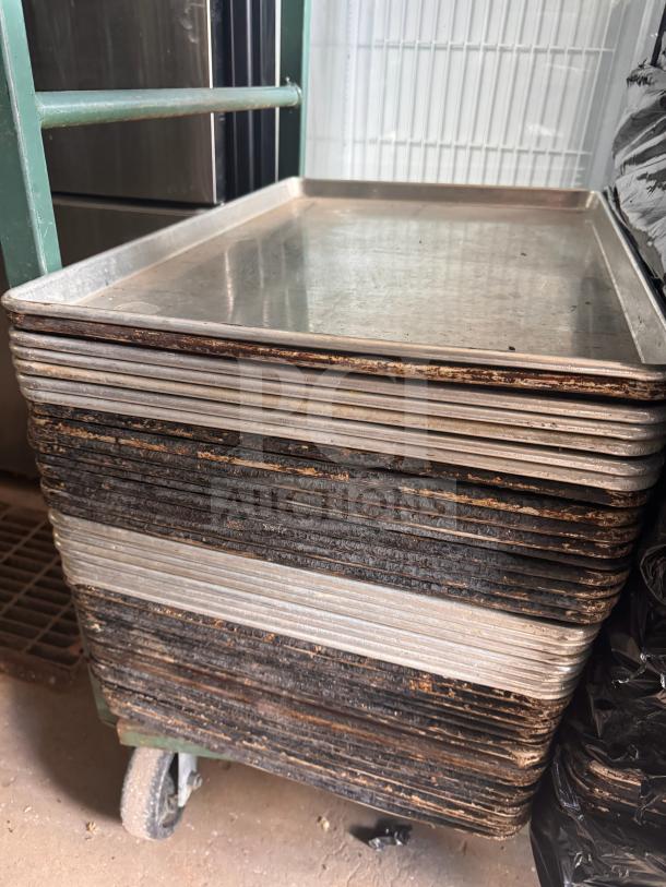 Stack of used stainless steel sheet pans on a green cart, showing signs of wear and patina from frequent use.