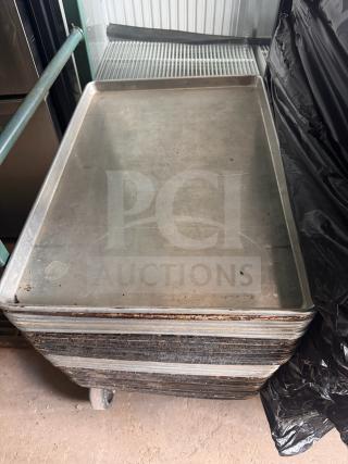 Stack of metal sheet pans, used condition, visible wear and discoloration, nested on a wheeled rack.