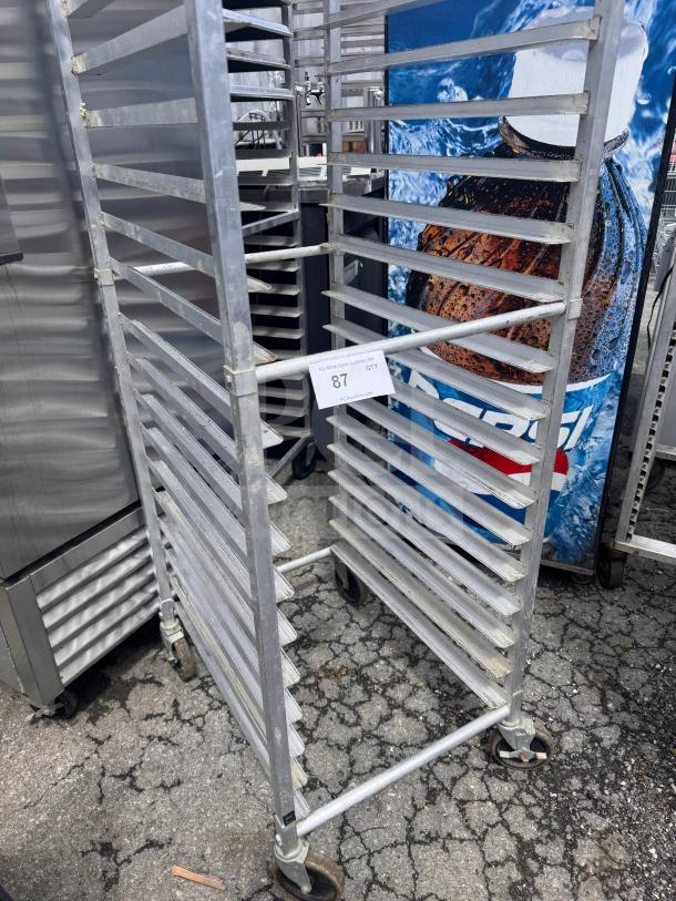 Steel sheet pan rack with multiple tiers, on wheels, moderate wear. Auction tag indicates lot 87. Ideal for bakery use.