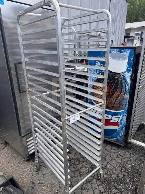Stainless steel sheet pan rack with multiple slots, caster wheels, and visible wear. Tag marked "88" hangs on the side.