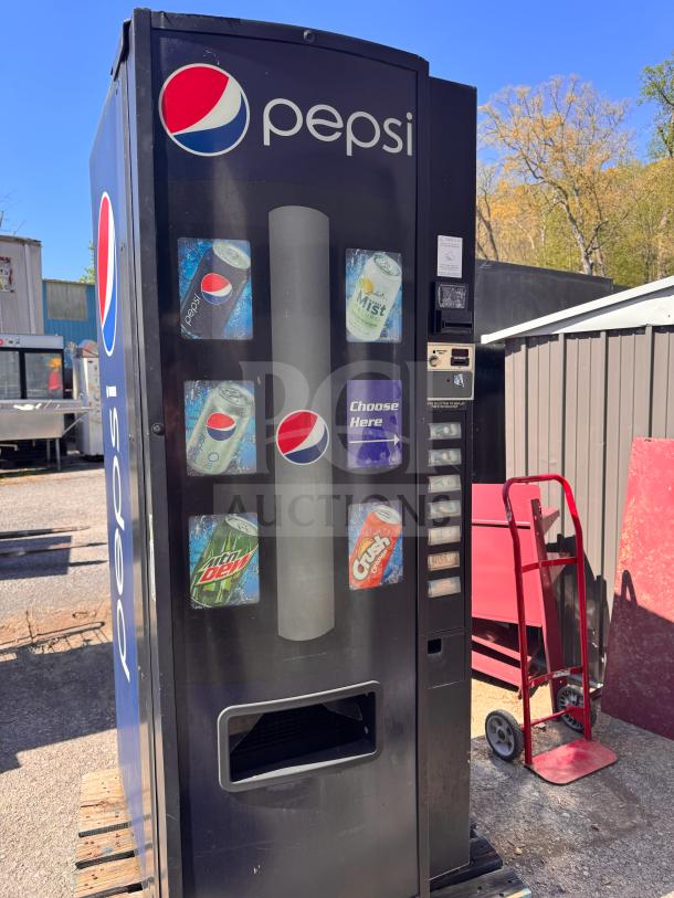 Pepsi vending machine displaying soda options, missing key, black exterior, coin slot visible, placed outdoors on a pallet.