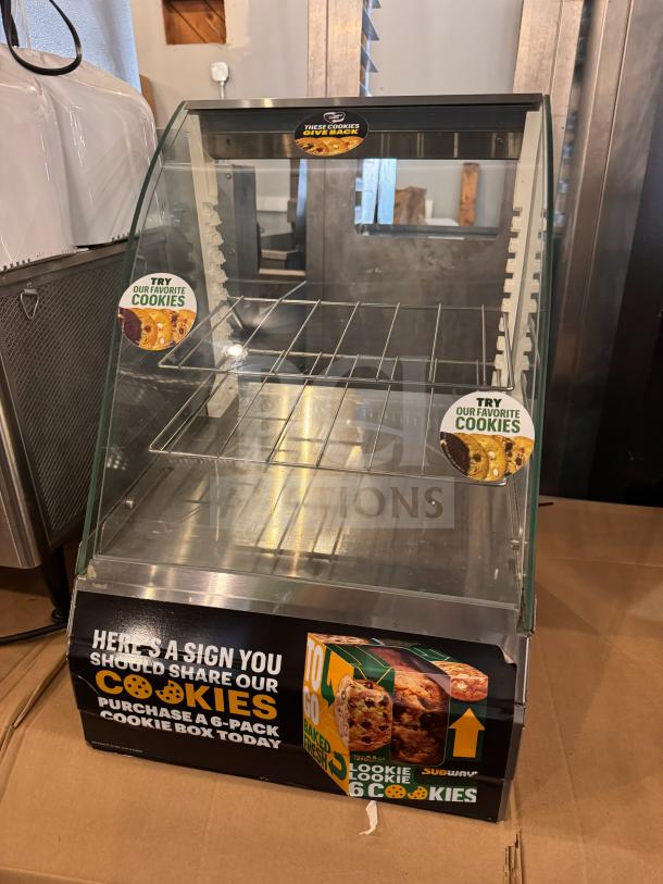Glass countertop display case for cookies with metal racks. Features promotional cookie labels and a "Purchase a 6-Pack" advertisement.