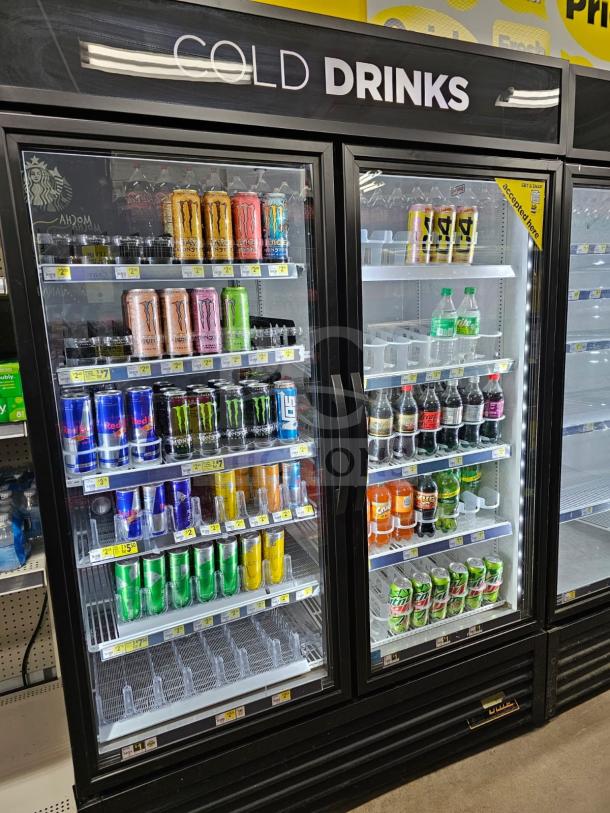 True two-door cooler merchandiser filled with beverages. Features clear glass doors, interior lighting, and labeled pricing. Working condition.
