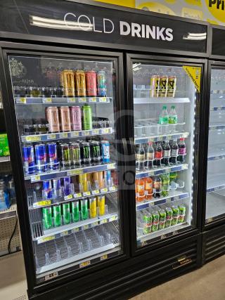 True two-door cooler merchandiser filled with beverages. Features clear glass doors, interior lighting, and labeled pricing. Working condition.