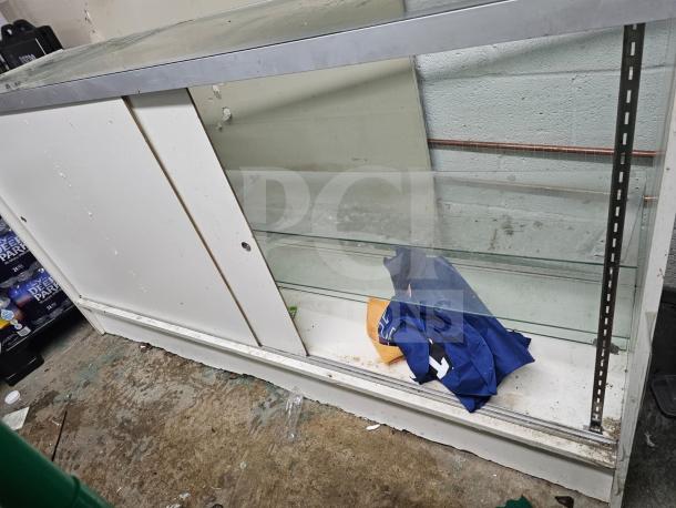 White display case with glass shelves and metal trim. Visible wear and dirt on the surface. Lower storage area behind sliding doors.