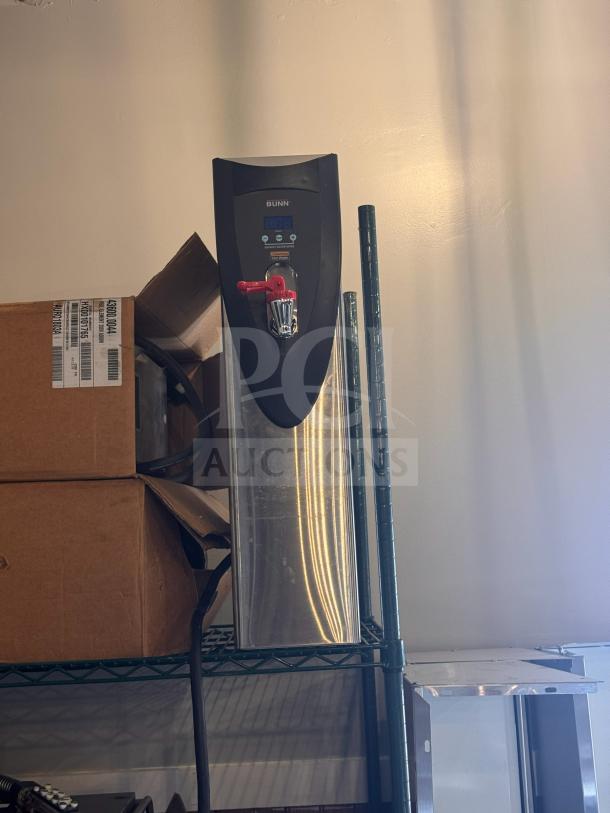 Brand new Bunn hot water dispenser in stainless steel, with digital display and red faucet, resting on a storage shelf.