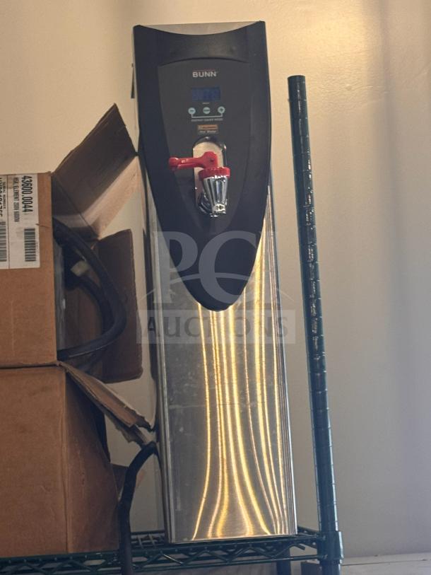 Brand new Bunn hot water dispenser with digital display and red tap, stainless steel body, on storage shelf.