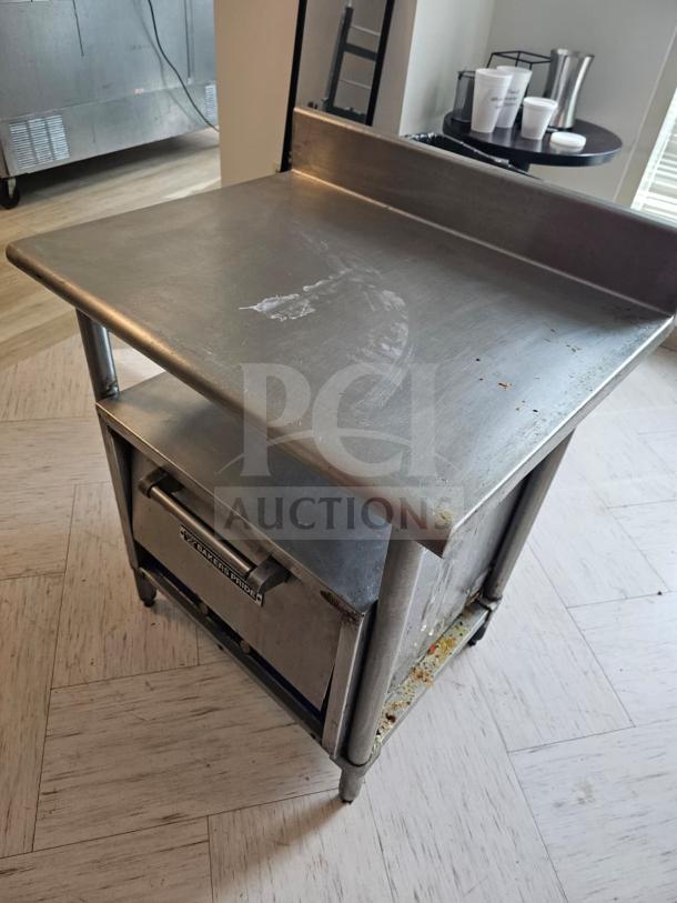 Stainless steel stand with backsplash, used condition. Includes lower shelf and a drawer labeled "Baker's Pride." Marks and wear visible.
